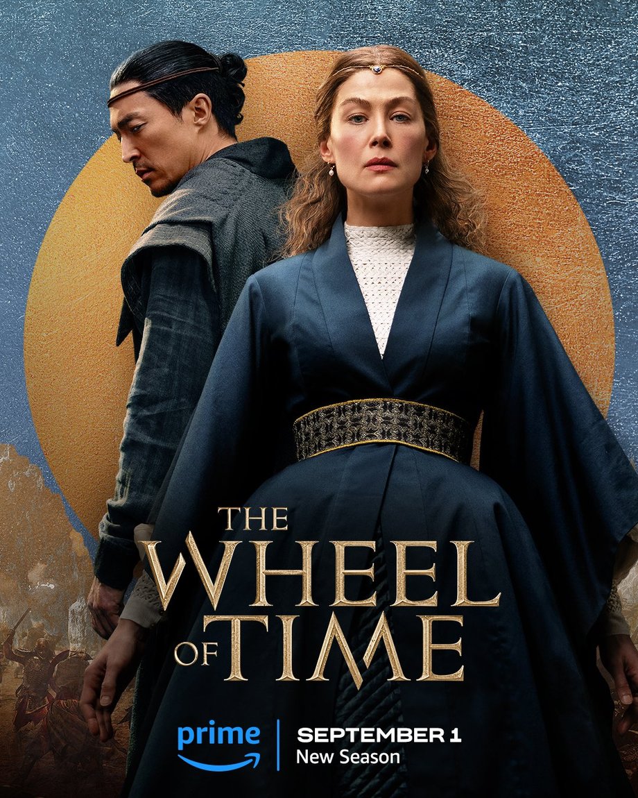 A large gold leaf-textured circle sits atop a blue foil background. The jutting peaks of mountains are beneath the circle, with people locked in battle in front of the range. Two characters are featured centered in the image. In the foreground on the right, Moiraine is in a medium shot. Her hair is worn half up, with a simple jeweled headpiece along her hairline and drop earrings; she wears a blue wrap dress; and her arms extend alongside the fuller skirt of her dress. Behind her to the left stands Lan, also shown in a medium shot. He stands with his back to us, looking over his left shoulder toward the lower left corner of the frame. Lan’s hair is pulled back in a bun with a leather band tied around his head; and he wears a blue vest and long-sleeve shirt. Golden text in the lower center reads, “The Wheel of Time.” At the bottom, the blue Prime Video logo sits next to white text that says “September 1, New Season.”