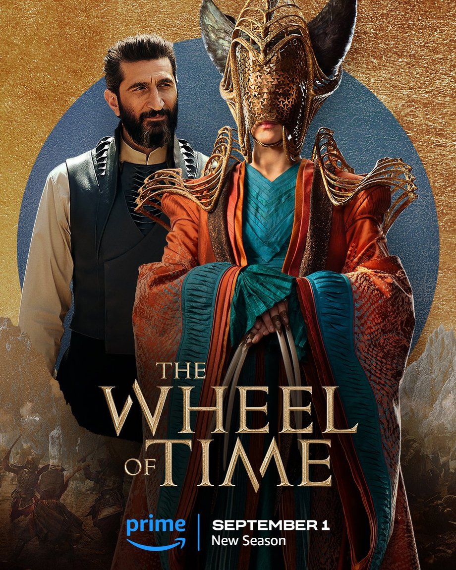 A blue foil circle sits atop a gold leaf-textured background. The jutting peaks of mountains are beneath the circle, with people locked in battle in front of the range. Two characters are featured centered in the image. In the foreground, High Lady Suroth is in a medium shot on the right side of the frame. She wears an elaborate teal gown and sunset colored coat with  embroidered sleeves; her face is covered by a large golden headpiece; structured golden accessories sit across her shoulders; and she has four extremely long nails. Behind her to the left stands Ishamael, also shown in a medium shot. His hair and beard are dark; he wears a white long-sleeve shirt with a collar; and a vest with woven detailing. Golden text in the lower center reads, “The Wheel of Time.” At the bottom, the blue Prime Video logo sits next to white text that says “September 1, New Season.”

