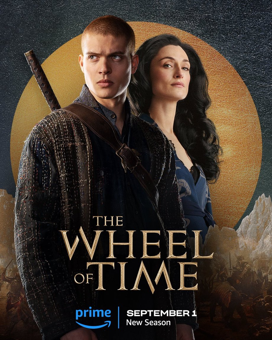 A large gold leaf-textured circle sits atop a slate-colored foil background. The jutting peaks of mountains are beneath the circle, with people locked in battle in front of the range. Two characters are featured centered in the image. In the foreground, Rand is featured in a medium shot. He wears a button-up shirt, a tweed-like coat, and his heron-marked blade on his back. Behind him stands Selene. Her dark curled hair cascades over her shoulders, she wears a gray-blue wrap dress, and she looks forward. Golden text in the lower center reads, “The Wheel of Time.” At the bottom, the blue Prime Video logo sits next to white text that says “September 1, New Season.”