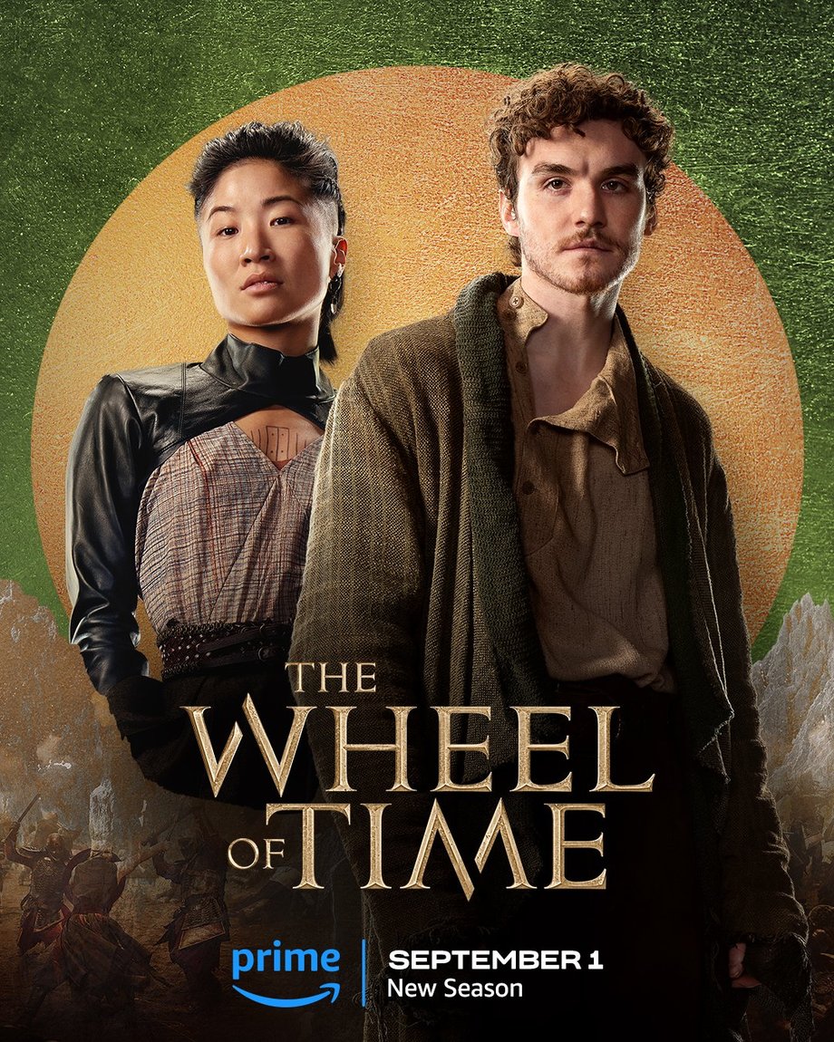 A large gold leaf-textured circle sits atop a green foil background. The jutting peaks of mountains are beneath the circle, with people locked in battle in front of the range. Two characters are featured centered in the image. In the foreground on the right, Mat is featured in a medium shot. He has curly short hair; wears a tan shirt; a tweed-like coat with an olive green shawl collar; and dark pants. He gazes forward. Behind him to the left stands Min, also shown in a medium shot. Her dark hair is pulled back; she wears a leather top and belt over a plaid v-neck; and she looks forward. Golden text in the lower center reads, “The Wheel of Time.” At the bottom, the blue Prime Video logo sits next to white text that says “September 1, New Season.”